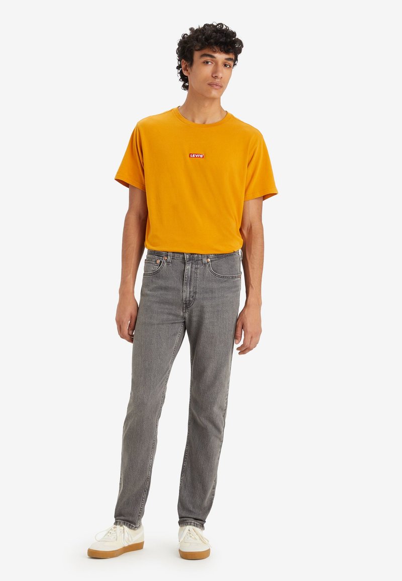 Tricou din bumbac galben cu un logo roșu Levi's, asortat cu blugi gri din denim și adidași albi cu talpă bej. Stil și croi casual.