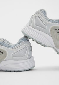 Light gray sneakers with mesh and synthetic materials, featuring textured accents, cushioned soles, and a modern, rounded design.