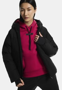 Fekete pufi dzseki egy élénk rózsaszín kapucnis pulcsi fölött. A dzsekin kapucni és cipzáras záródás található, míg a pulcsi zsinórral ellátott és logó részletekkel rendelkezik.