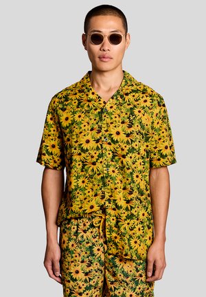 Mand iført matchende gult blomsterprint skjorte- og kortsæt med solbriller, stående mod en ensfarvet grå baggrund.