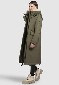 Lange olijfgroene parka met een met bont gevoerde capuchon, rits aan de voorkant, zijzakken en manchetten, gecombineerd met beige platformschoenen. Glad, duurzaam materiaal.