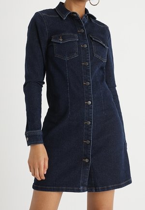 Mujer que lleva un vestido de mezclilla azul oscuro con botones, mangas largas, bolsillos con solapa en el pecho y cuello, de pie con una mano hacia abajo.