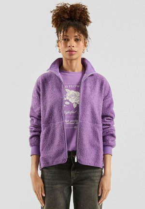 Kobieta o kręconych włosach, ubrana w fioletową bluzę polarową na zamek, pod którą ma koszulkę z nadrukiem, oraz ciemne dżinsy, stoi na tle jasnego, jednolitego tła.