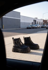 Bottes épaisses noires à plateforme avec double boucle, posées sur le trottoir près d’une rue avec des voitures garées et des bâtiments industriels en arrière-plan.