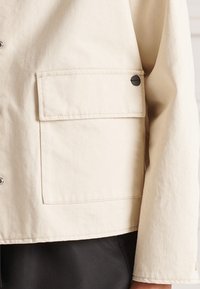 Beżowa kurtka z płótna z kieszenią na przedzie, zapięciem na napy i widocznymi detalami szwów. Zawiera czarny okrągły akcent na kieszeni.