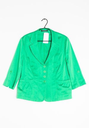 Blazer verde da donna con maniche tre quarti, quattro bottoni, due tasche frontali, appeso su una gruccia nera con etichetta bianca.