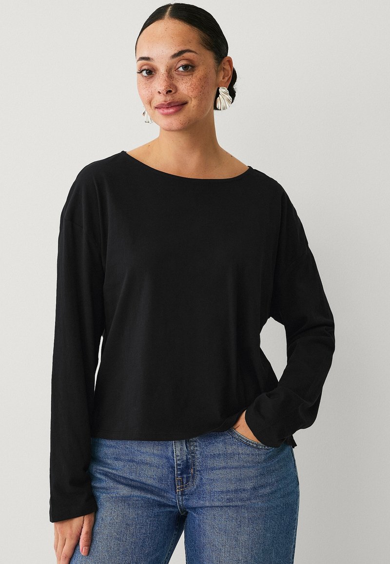 Maglietta nera a maniche lunghe realizzata in materiale morbido, con scollatura ampia, vestibilità comoda e cuciture in vista. Abbinata a jeans in denim blu.