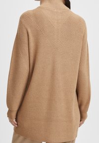 Maglione beige a maglia con colletto alto, caratterizzato da un motivo testurizzato e dettagli di cucitura a V sulla schiena. Maniche lunghe e larghe.