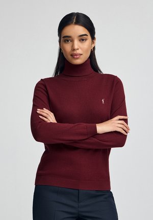 Femme aux cheveux bruns, portant un pull col roulé bordeaux et un pantalon bleu marine, se tenant les bras croisés sur un fond clair et uni.