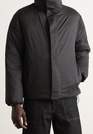 Schwarze wattierte Jacke mit hohem Kragen und verdecktem Frontreißverschluss, getragen zu dunklen Jeans aus Denim, Hand teilweise sichtbar in der Tasche.