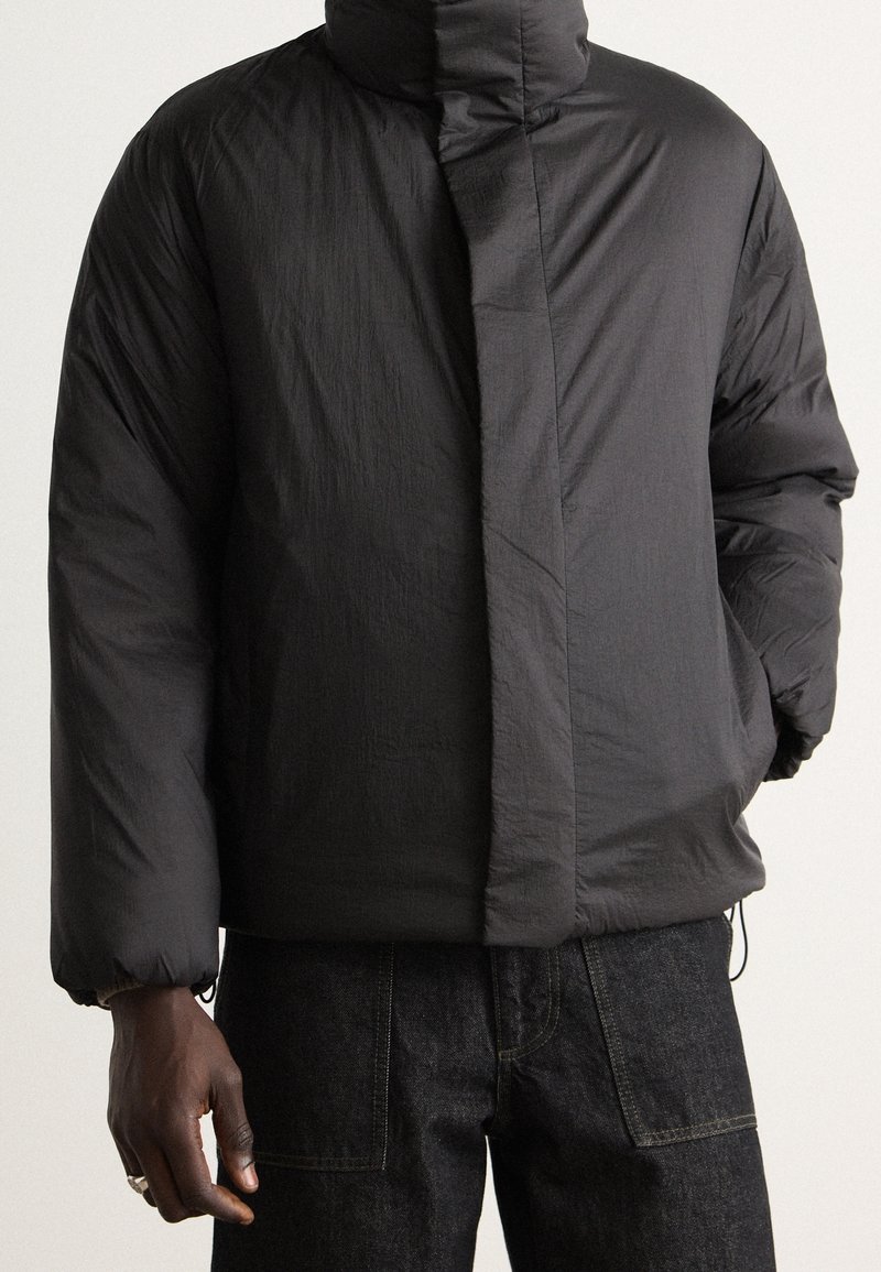Veste matelassée noire avec col montant et fermeture éclair avant dissimulée, portée avec un jean en denim foncé, main partiellement visible dans la poche.