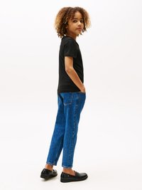T-shirt noir à manches courtes, jeans en denim bleu avec une coupe décontractée et chaussures noires. Cheveux bouclés, longueur épaule. Debout avec les mains dans les poches.