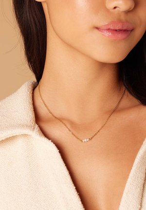 Gouden ketting met drie kleine, ronde witte kralen, gedragen op een blootgestelde halslijn, met een zachte, textuurachtige stofkraag zichtbaar.