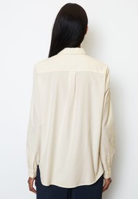 Camisa de manga larga beige clara con cuello, corte relajado, aberturas laterales y puños abotonados. Tejido suave con textura mínima.