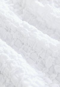 Tessuto bianco con texture e volant arricciati, che presenta un motivo a griglia. Il materiale appare morbido e leggero, sottolineando un design delicato.