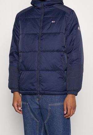 Man draagt een marineblauwe doorgestikte jas met rits aan de voorkant en zijzakken, gecombineerd met blauwe spijkerbroek, staand met ontspannen armen.