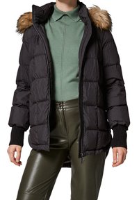 Mujer vestida con una chaqueta acolchada negra con capucha de piel sintética, suéter de punto verde y pantalones de cuero verde oliva, de pie con las manos relajadas.