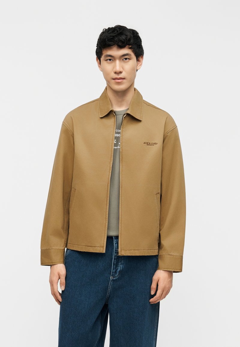 Νέος άνδρας με κοντά μαύρα μαλλιά που φοράει μια καφέ ζακέτα με φερμουάρ πάνω από γκρι πουκάμισο και μπλε τζιν, όρθιος μπροστά σε ένα απλό φόντο.