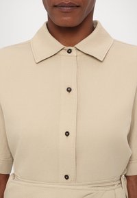 Gros plan d'une personne portant une chemise beige à boutons, col pointu et manches courtes, montrant le haut du torse et le détail de la ceinture à la taille.