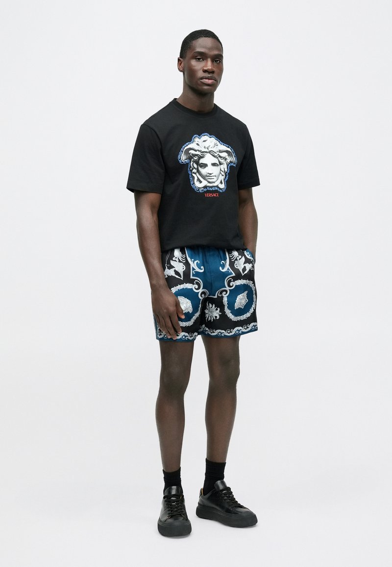 Black t-shirt with a graphic of a face, paired with patterned blue and black shorts. Model wears black sneakers and socks.