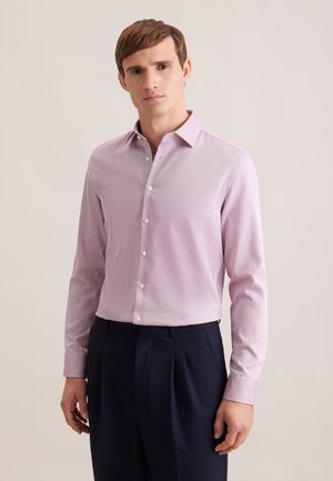 Uomo con camicia rosa chiaro a maniche lunghe con bottoni e pantaloni plissettati blu scuro, in piedi davanti a uno sfondo beige semplice.