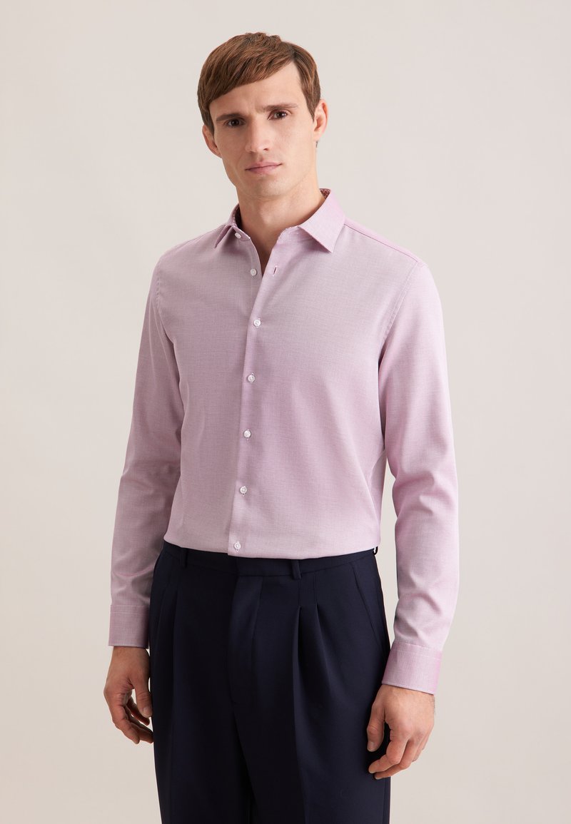 Homme portant une chemise à manches longues rose clair boutonnée et un pantalon plissé bleu marine foncé, debout devant un fond beige uni.