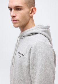 Άνδρας με κοντά μαλλιά και σκουλαρίκια που φοράει ανοιχτό γκρι φούτερ Björn Borg, κοιτάζοντας ελαφρώς προς τα κάτω και αριστερά, μπροστά σε απλό φόντο.