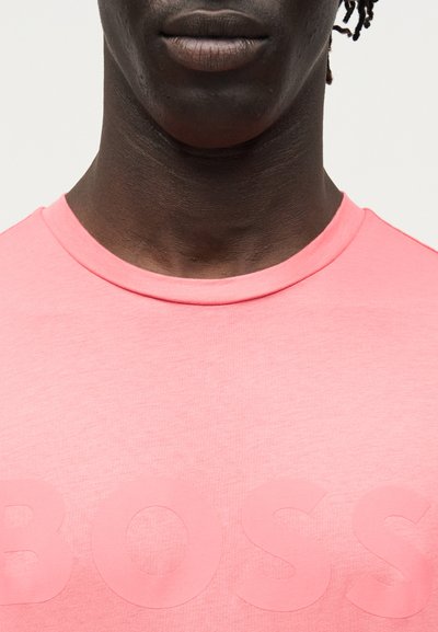 Gros plan sur la partie inférieure du visage et le cou d’une personne portant un t-shirt rose à col rond avec des lettres en relief formant le mot « BOSS ».