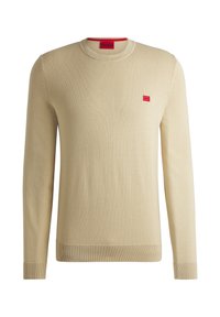 Pull en maille beige avec col rond, manches longues, poignets et ourlet côtelés, présentant une petite étiquette logo rouge sur la poitrine.