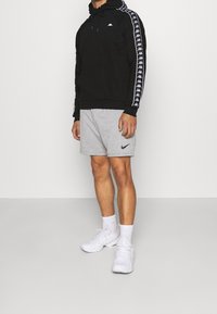 Felpa nera con cappuccio, logo bianco e strisce laterali, abbinata a pantaloncini grigi con logo nero e sneakers bianche.