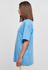 Femme aux cheveux bruns et raides portant un t-shirt ample à manches courtes bleu clair, debout de profil sur un fond uni.