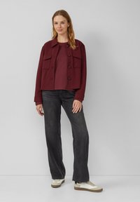 Bordeauxrot cropped Jacke mit Knopfschluss und zwei vorderen Taschen, kombiniert mit einem passenden Oberteil, schwarzen weiten Jeans und beigen Sneakers.