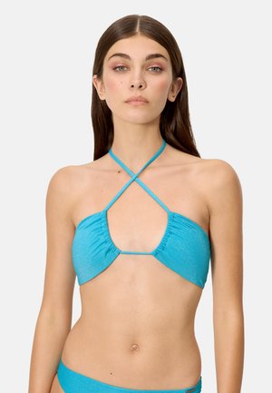 Mujer con cabello largo y castaño vistiendo un top de bikini azul turquesa brillante con tirantes cruzados en el cuello, sobre un fondo liso.