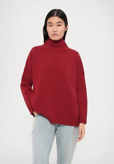 Un pull à col roulé rouge, épais, avec une coupe ample, des accents côtel�és au niveau du col et des manches, associé à un jean bleu clair.