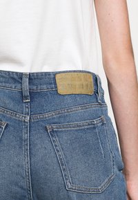 Närbild av baksidan av en person som bär blå denimjeans med ett ljust märke på midjebandet. En enkel vit skjorta syns ovanför.