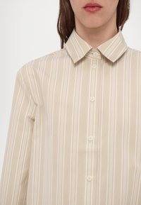 Beige stripete skjorte med knappekrage, med hvite vertikale striper og lyse knapper, laget av glatt stoff.