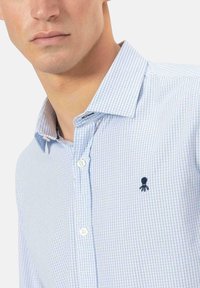 Camisa de cuadros azul claro hecha de algodón, con cuello abotonado y un pequeño logo de pulpo bordado en el pecho.