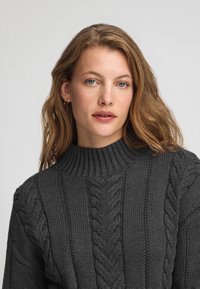Ciemnoszary dziergany sweter z wysokim prążkowanym kołnierzem, zdobiony wzorami warkoczowymi i teksturowanymi detalami na rękawach.
