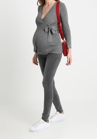 Top de maternidad gris con canalé y un detalle de lazo en la cintura, combinado con jeans pitillo grises y zapatillas blancas. Bolso de hombro rojo como acento.