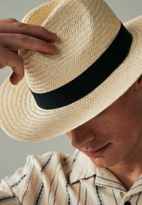 Chapeau en paille à large bord avec une texture tissée. Présente un bandeau en tissu noir autour de la base. Le chapeau est légèrement incliné.