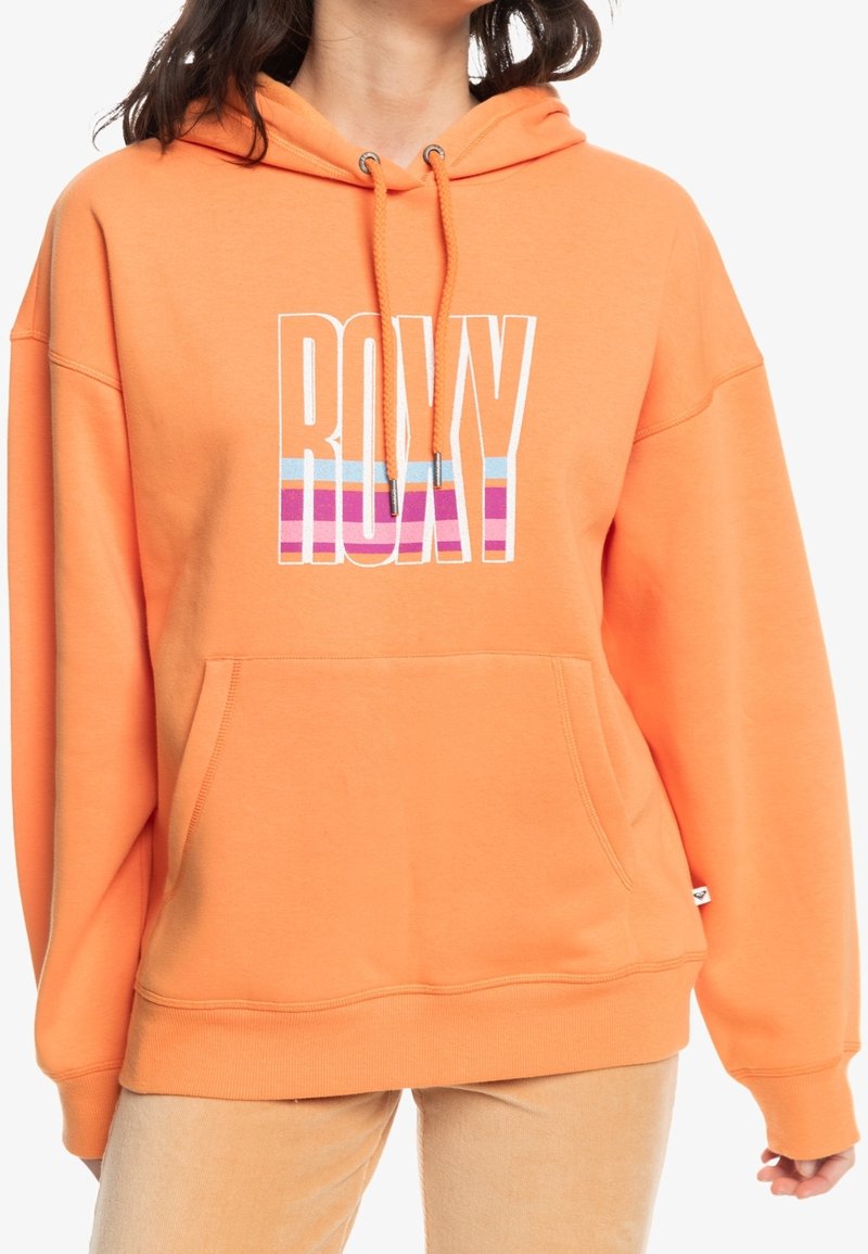Personne portant un sweat à capuche orange oversize avec le texte "ROXY" et des rayures colorées à l'avant, associé à un pantalon beige clair.