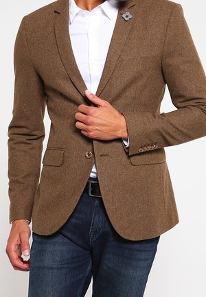 Hombre vestido con blazer marrón con pin de flor en la solapa, camisa blanca, vaqueros oscuros, ajustando el botón del blazer con una mano.