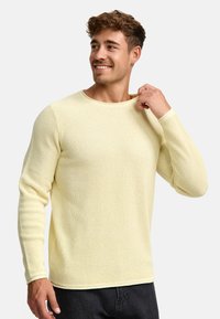 Maglione a maniche lunghe in maglia a nido d'ape gialla con collo rotondo e polsini aderenti. Il modello sta tirando il colletto, indossando pantaloni scuri.