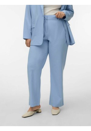 Kvinne iført lyseblå skreddersydde bukser og matchende blazer, beige lave slip-on sko, stående på grått underlag.