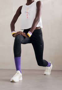 Valkoinen lyhyt tankkitoppi, jossa on punainen ja harmaa grafiikka; mustat kuviolliset legginsit; violetit, valkoiset ja keltaiset ranne- ja pohjevahvikkeet; valkoiset urheilukengät.