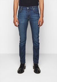 Jeans in denim blu slim fit con texture usurata, design a cinque tasche e cuciture a contrasto. Abbinati a scarpe nere stringate.