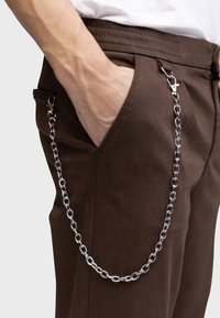 Pantalons marron avec un accessoire en chaîne. La chaîne est composée de maillons en métal argenté, attachée à une passant de ceinture avec un fermoir. Tissu texturé.