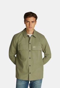 Camicia verde oliva in cotone con taglio rilassato, dotata di due tasche sul petto e un piccolo logo sulla tasca sinistra.