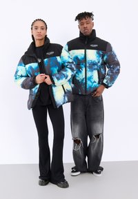 Pufferjakker med sorte og blå abstrakte mønstre, kontrasterende sorte skuldre og lynlåse; modellen er parret med flagrende bukser og slidte jeans.