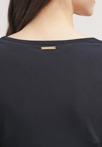 Žena oblečená v černém topu s malou zlatou destičkou logo Michael Kors umístěnou uprostřed pod výstřihem vzadu.